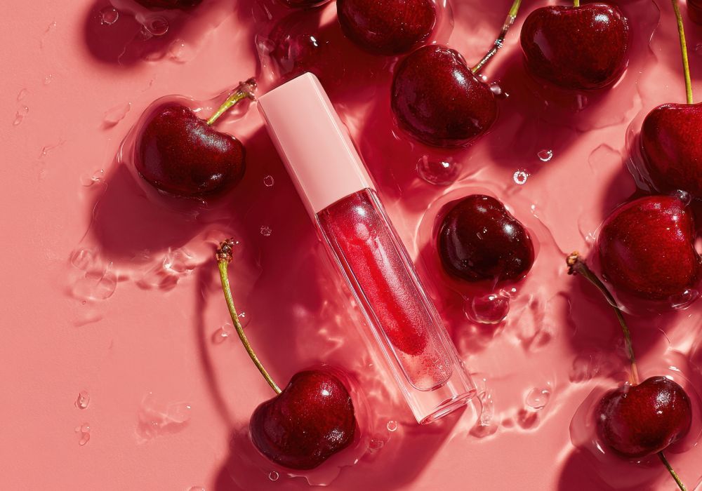 Gloss rose dans un tube transparent entouré de cerises fraîches, évoquant un maquillage maison aux notes fruitées.
