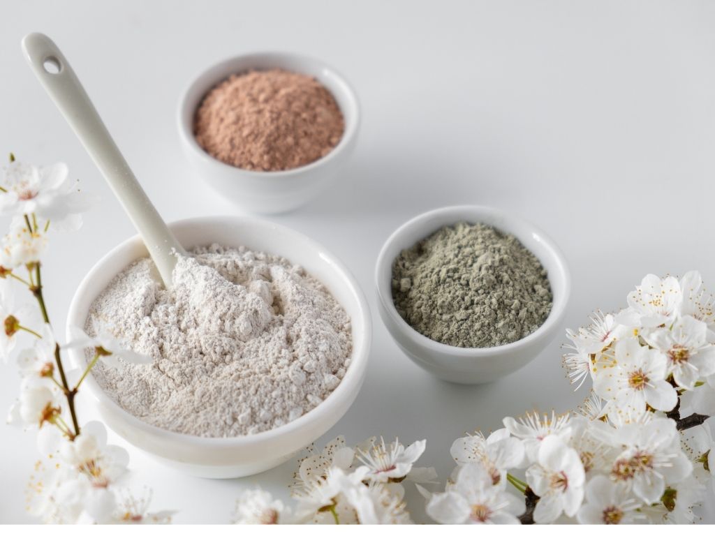 Trois bols d'argile cosmétique, rose, blanche et verte, entourés de fleurs