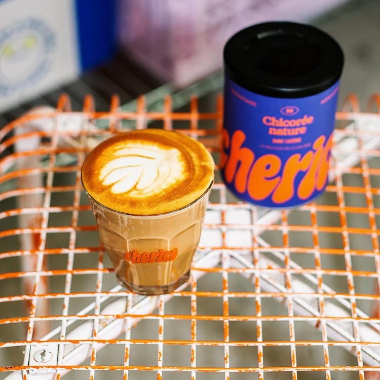 Un verre siglé Cherico rempli de chicorée au lait coiffée d'un dessin en mousse, posé sur une table de terrasse en grillage orange, à côté d'un pot bleu de Chicorée nature sans caféine