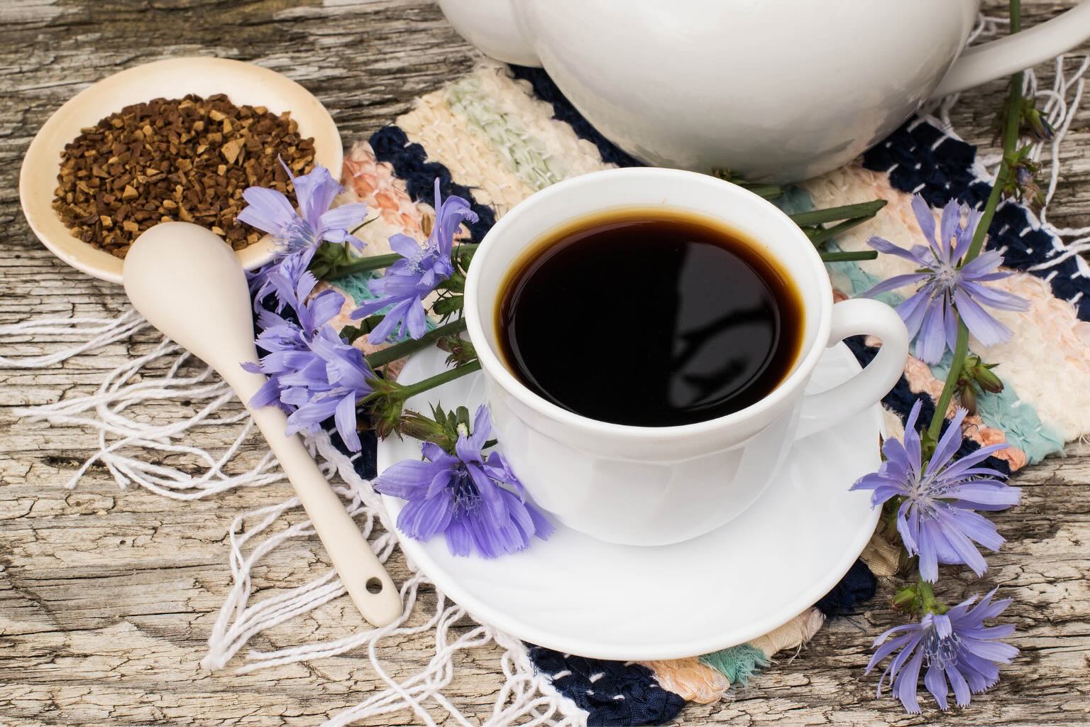 Une tasse de chicorée noire sur sa soucoupe, à côté d'une coupelle de chicorée en grains concassés et d'une cuillère en céramique, avec des fleurs de chicorée bleues posées sur un napperon tissé et une table en bois usé