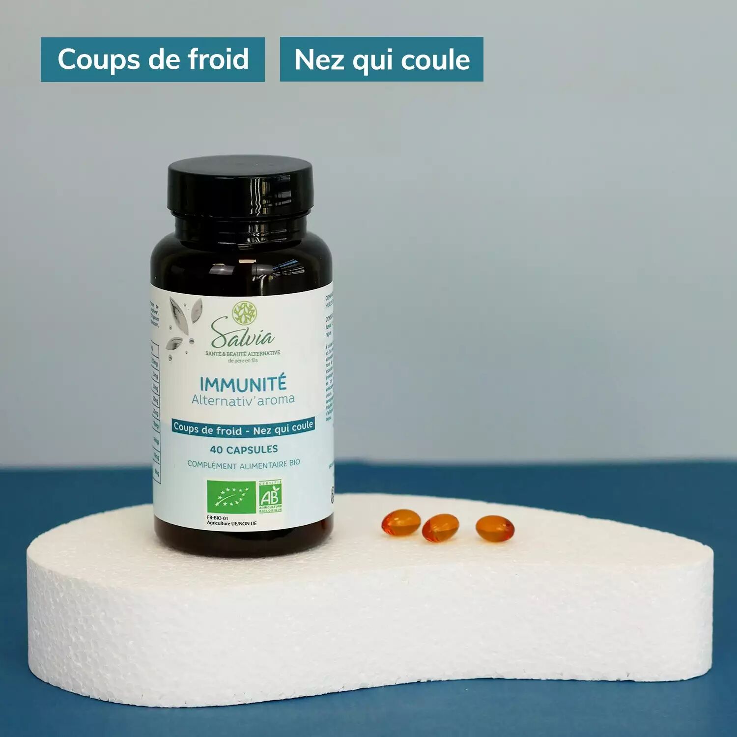 Pot d'Alternativ'aroma de Salvia, 40 capsules bio, posé sur un socle blanc à côté de trois capsules ambrées, sur fond bleu, avec les étiquettes de la marque « Coups de froid » et « Nez qui coule »