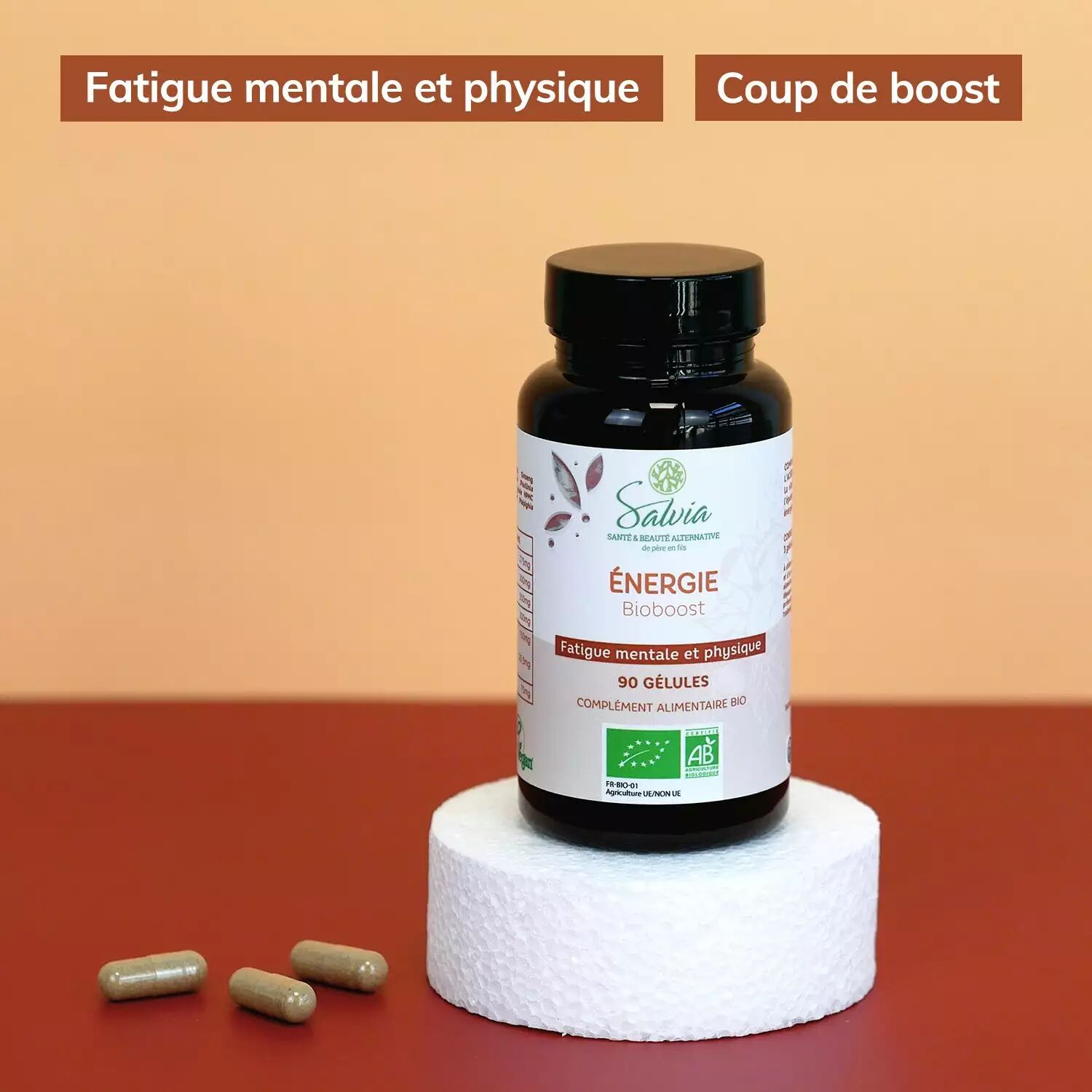 Pot de Bioboost de Salvia, 90 gélules bio, sur un socle blanc devant trois gélules beiges, sur fond orangé, avec les étiquettes de la marque « Fatigue mentale et physique » et « Coup de boost »
