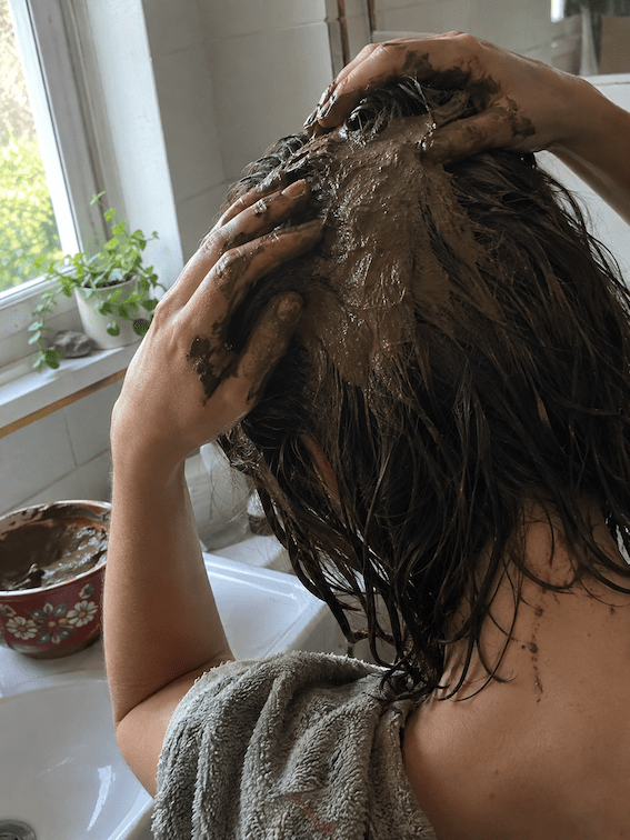 Vue de dos d'une personne aux cheveux bruns mouillés qui masse une pâte de rhassoul brune sur son cuir chevelu à deux mains, avec des coulures dans le cou, une serviette sur l'épaule et un bol de pâte posé au bord de la baignoire