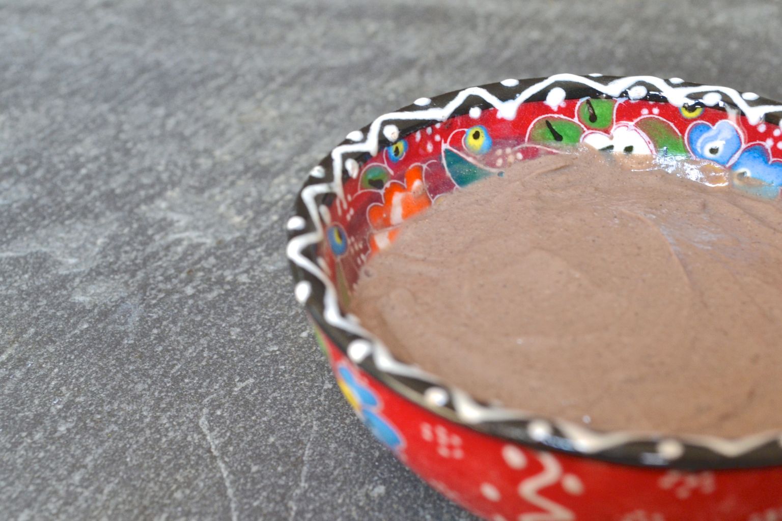 Pâte de rhassoul brune et lisse, prête à appliquer, dans un bol en céramique peinte à fleurs posé sur un plan de travail gris