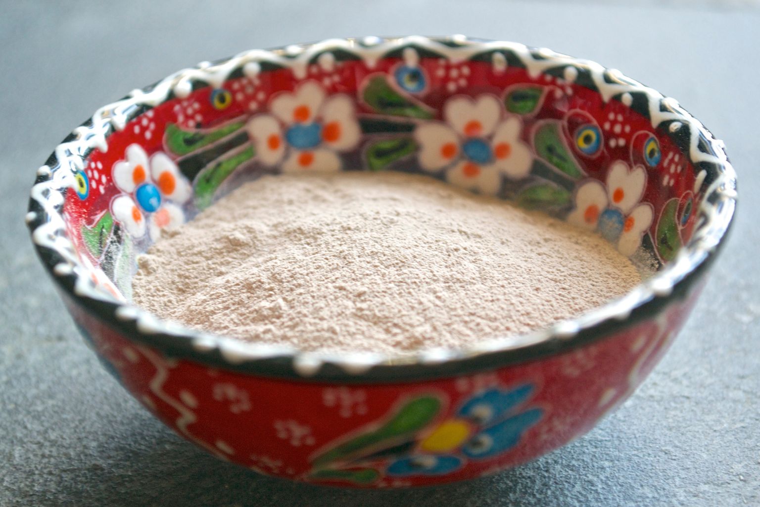 Poudre de rhassoul beige clair dans un bol en céramique rouge peinte de fleurs blanches et bleues, sur fond gris